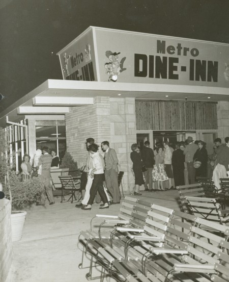 Black and white photograph of men and women lining up to enter a building called the ‘Metro Dine-Inn’. The building is surrounded by patio chairs and pot plants. 